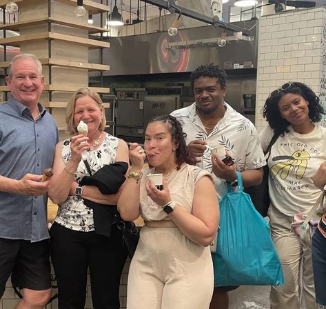 Five adults smiling and sampling ice cream at an urban ice cream shop counter with industrial kitchen equipment, subway-tile wall, hanging bulbs and shelving — one with a cone, others with cups and a bright blue shopping bag.