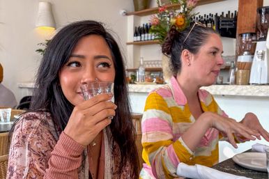 Two women chatting at a bright café table — one smiling and sipping from a glass, the other gesturing; marble counter, wine bottles and flowers in the background.