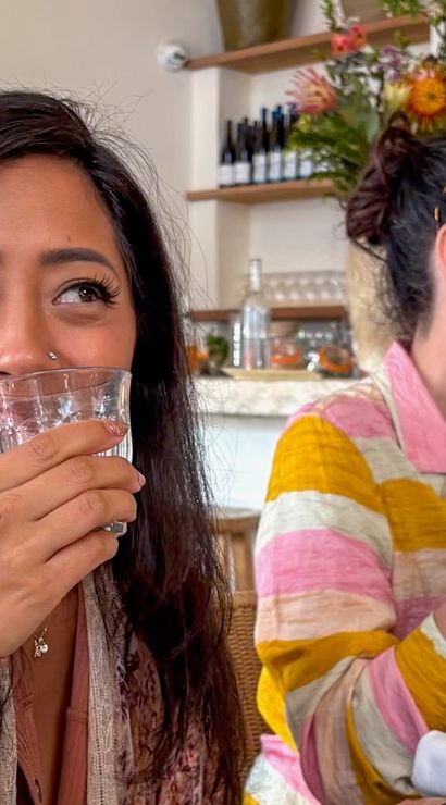 Two women chatting at a bright café table — one smiling and sipping from a glass, the other gesturing; marble counter, wine bottles and flowers in the background.