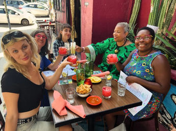 Five women smiling and toasting pink cocktails at a colorful sidewalk patio restaurant with chips, salsa, menus and tall cacti against a red wall.
