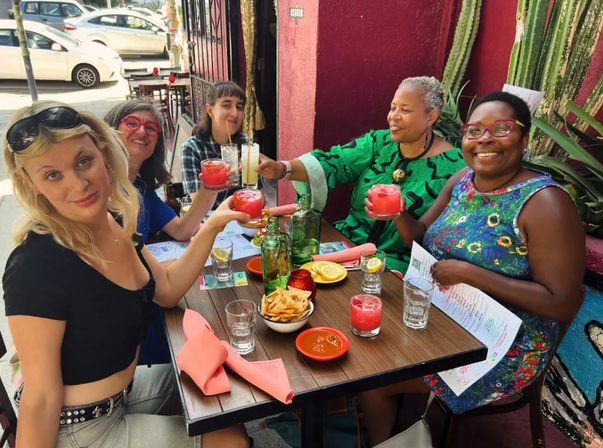 Five women smiling and toasting pink cocktails at a colorful sidewalk patio restaurant with chips, salsa, menus and tall cacti against a red wall.