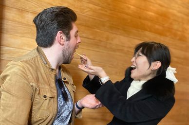 Playful moment of a laughing woman in a black coat feeding a man in a tan jacket with chopsticks against a warm wood-paneled interior backdrop