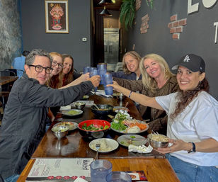 Cheerful group of six smiling friends clinking blue plastic cups over bowls of Asian-style dishes on a long wooden table inside a cozy modern restaurant.