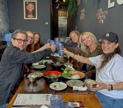 Cheerful group of six smiling friends clinking blue plastic cups over bowls of Asian-style dishes on a long wooden table inside a cozy modern restaurant.