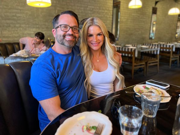 Smiling pair dining in a cozy brick-walled restaurant booth, plates and water glasses on a glossy table with a smartphone, under warm pendant lights.