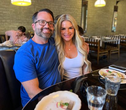 Smiling pair dining in a cozy brick-walled restaurant booth, plates and water glasses on a glossy table with a smartphone, under warm pendant lights.