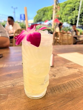 Tropical pale-yellow cocktail in a tiki glass with crushed ice, bamboo straw and purple orchid garnish on a wooden table at a sunny outdoor café patio