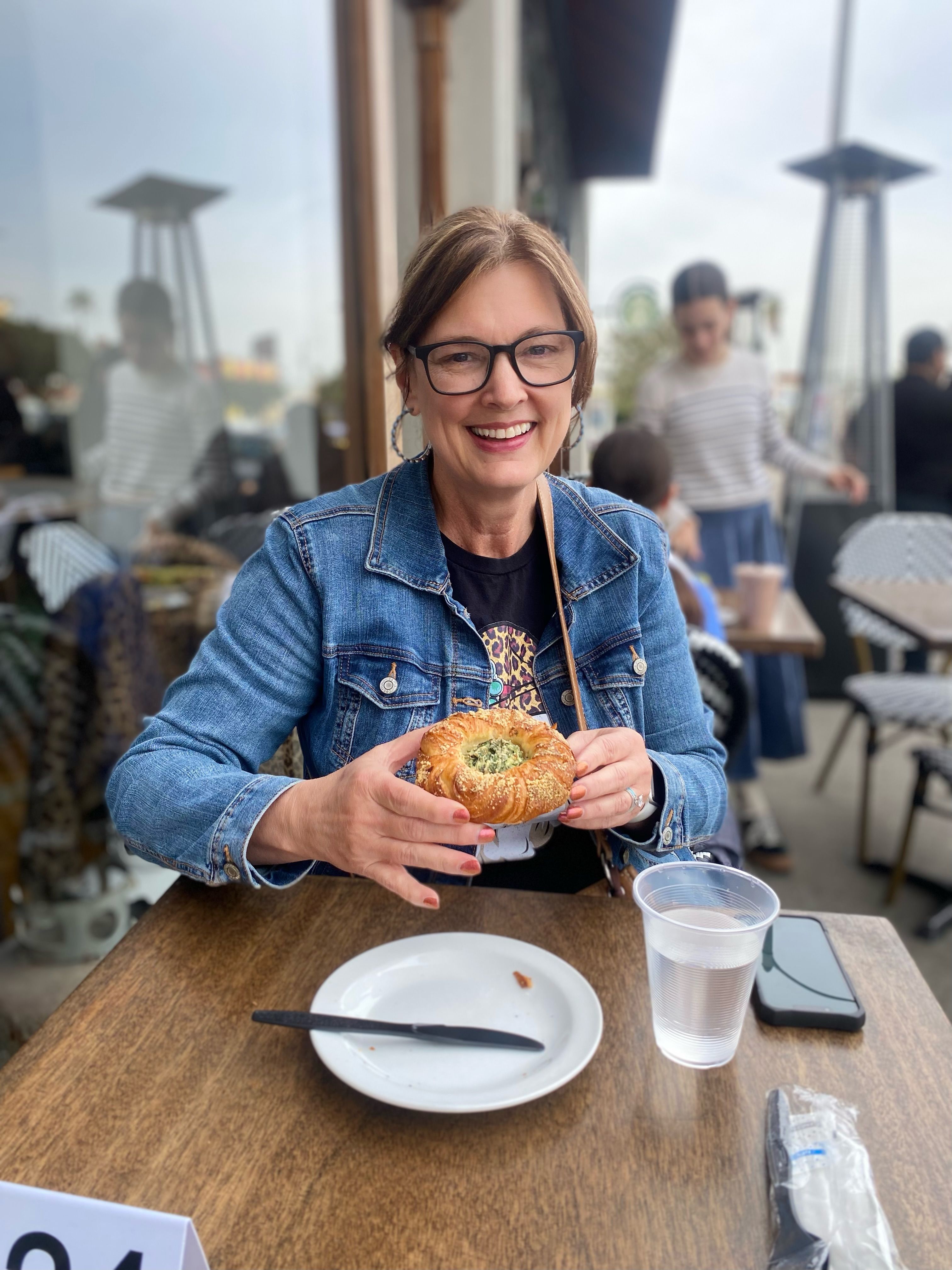 Smiling woman in a denim jacket and glasses at an outdoor café patio holding a flaky herb-filled pastry, with a plate, plastic cup of water and smartphone on the table — casual brunch scene.