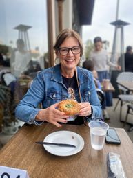 Smiling woman in a denim jacket and glasses at an outdoor café patio holding a flaky herb-filled pastry, with a plate, plastic cup of water and smartphone on the table — casual brunch scene.