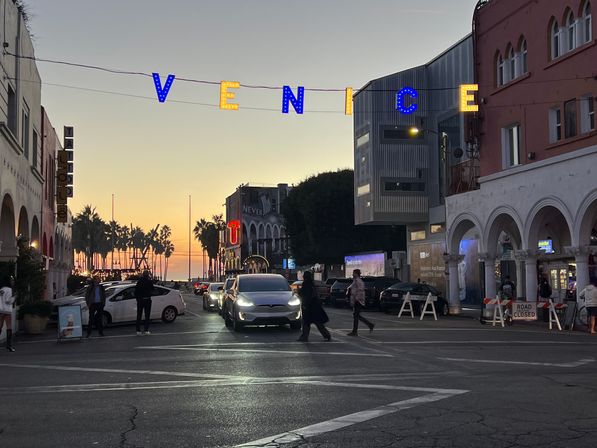 Sunset street scene at Venice Beach, California with illuminated 'VENICE' sign overhead, palm trees, pedestrians crossing, cars and arched storefronts leading to the ocean glow.