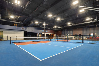 Spacious indoor pickleball courts with blue and orange playing surfaces, low nets and chain-link dividers beneath a high industrial ceiling with bright overhead lights and a viewing/snack area.