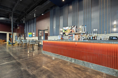 Sleek modern hotel bar and lounge with a long red-tiled counter and marble base, backlit shelves of bottles and beer taps, wall-mounted TV, high stools and casual seating on polished concrete under exposed high ceilings.