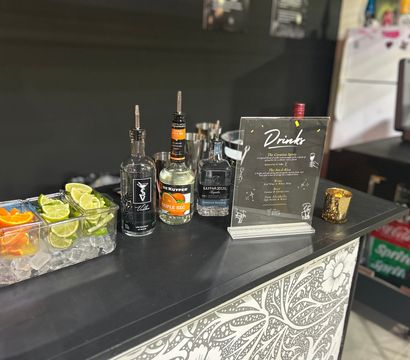 Cocktail bar station on a black counter with bottles of vodka, triple sec and tequila, clear trays of lime and orange wedges on ice, and a framed drinks menu.