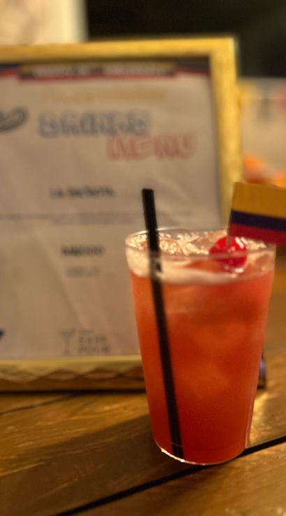 Bright red tropical cocktail with a cherry and small Colombian flag garnish on a wooden table, blurred framed menu in the background