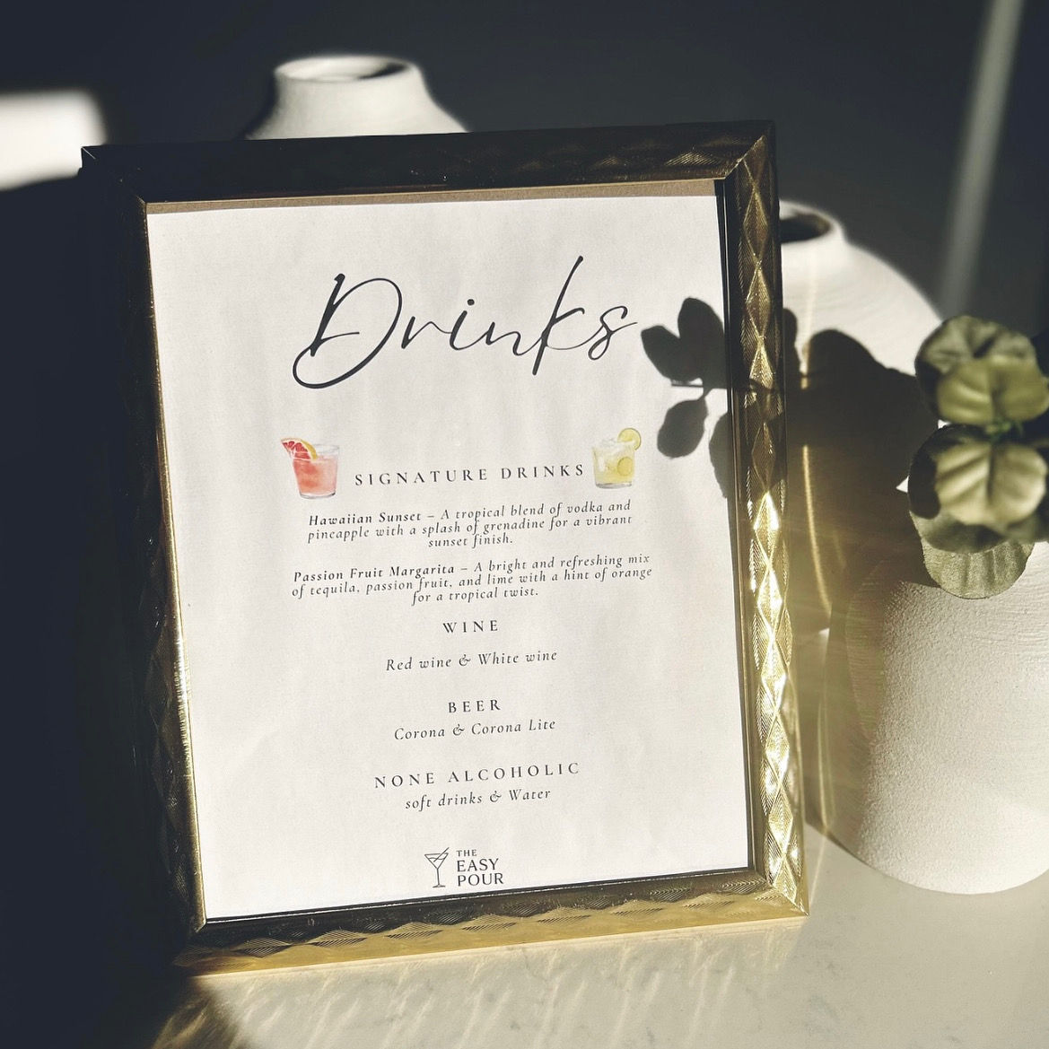 Sunlit gold-framed drinks menu on a tabletop with white vases and a potted plant shadow, listing signature cocktails, wine, beer and non-alcoholic options.