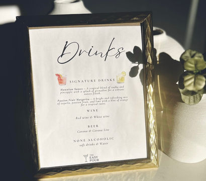 Sunlit gold-framed drinks menu on a tabletop with white vases and a potted plant shadow, listing signature cocktails, wine, beer and non-alcoholic options.