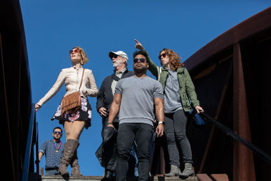 Five adults walking down metal bridge stairs on a sunny blue-sky day — a woman in a floral skirt and fringe bag, a man in a gray T‑shirt leading, and others in jackets and sunglasses, casual outdoor scene.