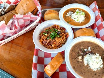 Colorful Southern spread on a wooden table: bowls of jambalaya topped with green onions, red beans and rice, a hearty gumbo with rice, soft rolls and wrapped sandwiches on red-and-white checkered paper — New Orleans-style Cajun/Creole comfort food.