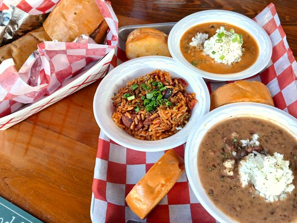 Colorful Southern spread on a wooden table: bowls of jambalaya topped with green onions, red beans and rice, a hearty gumbo with rice, soft rolls and wrapped sandwiches on red-and-white checkered paper — New Orleans-style Cajun/Creole comfort food.