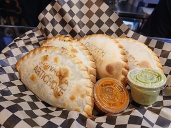 Golden-brown baked empanadas stamped 'ARTICHOKE' and 'CHICKEN' on black-and-white checkered deli paper with two dipping sauces, orange spicy and green herb.