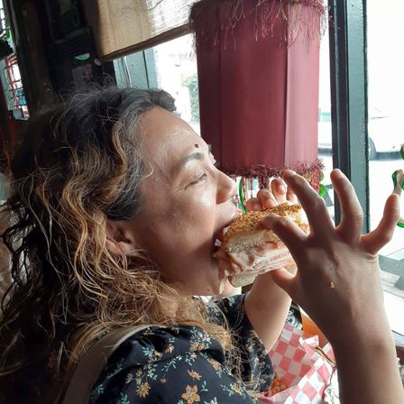 Person with curly hair taking a big bite of a stacked deli sandwich with sesame seed bun at a sunny café window, checkered paper-lined basket and drink nearby.