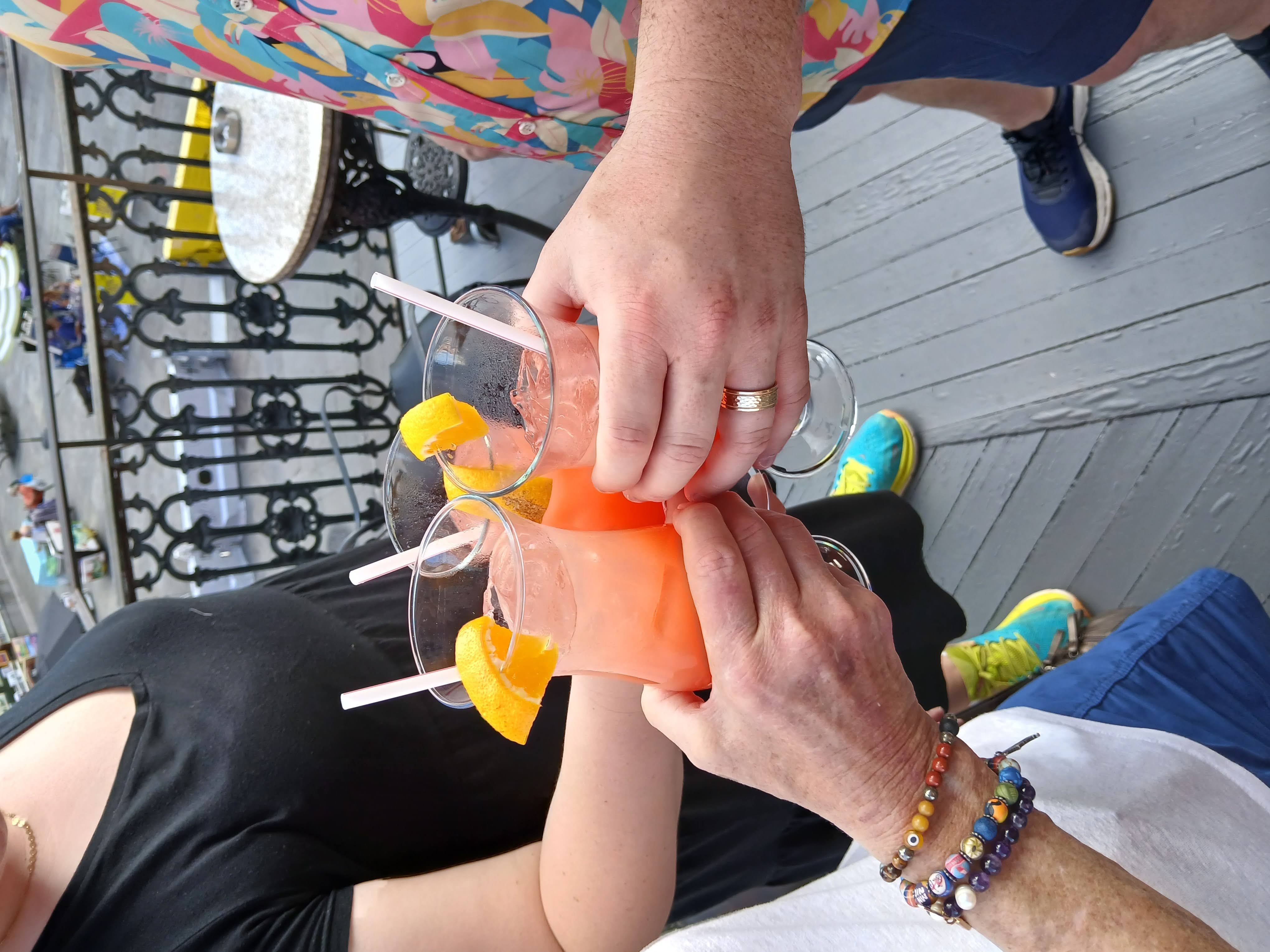 Three people toasting pink citrus cocktails garnished with orange wedges on a rooftop balcony patio overlooking a city street, colorful summer outfits visible.