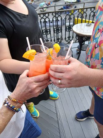 Three people toasting pink citrus cocktails garnished with orange wedges on a rooftop balcony patio overlooking a city street, colorful summer outfits visible.