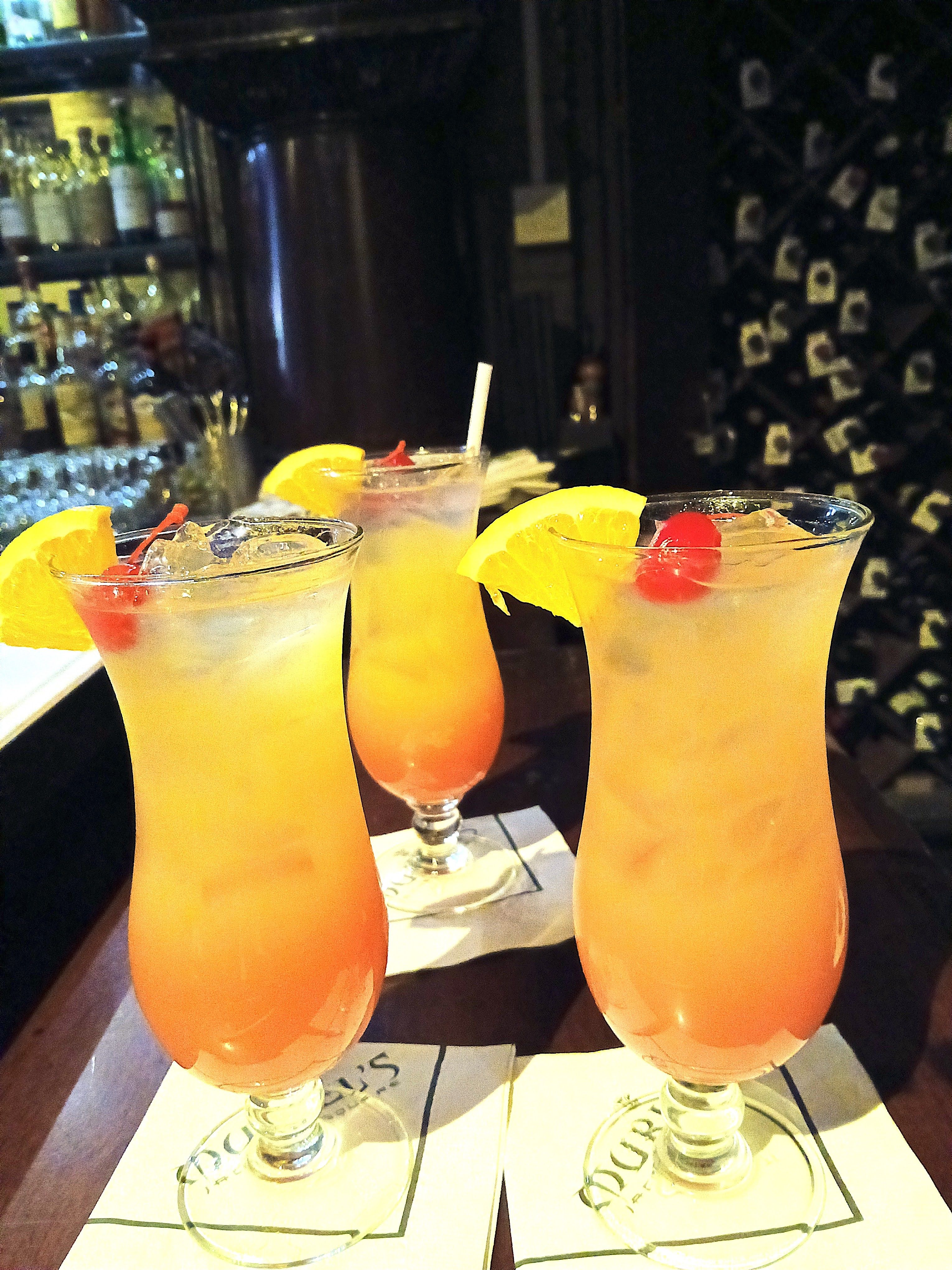 Three vibrant orange-pink tropical cocktails in hurricane glasses, garnished with orange slices and maraschino cherries, sitting on a bar counter with bottles and a wine rack in the background.
