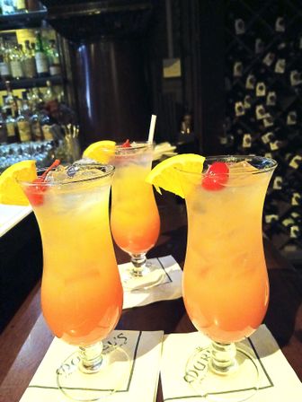 Three vibrant orange-pink tropical cocktails in hurricane glasses, garnished with orange slices and maraschino cherries, sitting on a bar counter with bottles and a wine rack in the background.