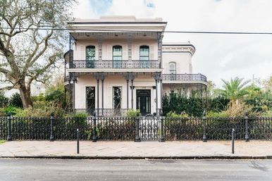 Pastel two-story New Orleans–style mansion with ornate wrought-iron balconies and fence, leafy garden, oak and palm trees, and a sidewalk-lined street in front.