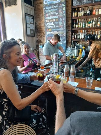 Group toasting lemon‑garnished cocktails at a lively rustic bar with exposed brick, a chalkboard drink menu and bartenders working behind the counter
