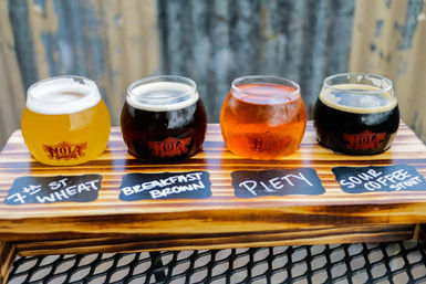 Craft beer flight on a wooden paddle holding four round tasting glasses—pale wheat, dark brown ale, amber ale, and a sour coffee stout—resting on a metal patio table.