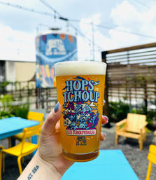 Hand holding a colorful illustrated pint of hazy craft beer with a frothy head in a sunny outdoor beer-garden patio in New Orleans, bright blue tables and yellow chairs blurred in the background.