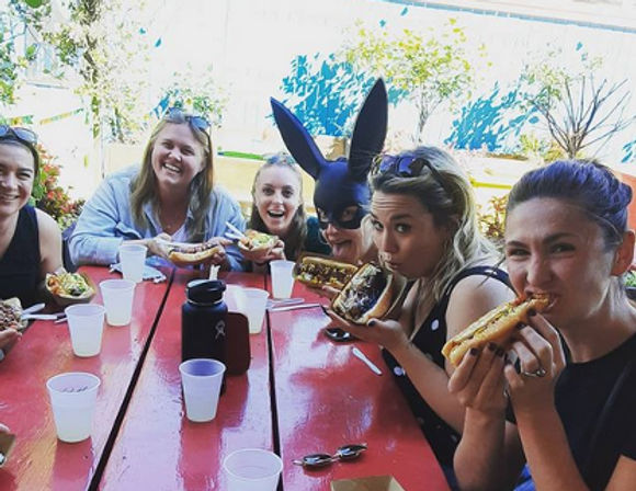 Group of friends at an outdoor red picnic table enjoying loaded hot dogs and drinks, one person wearing a playful black rabbit mask — casual summer meal.