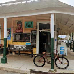 Yellow neighborhood corner store with covered porch, wooden bench, colorful hand-painted signs, chalkboard specials and an ice cooler, a red vintage bicycle locked to a green post in front.