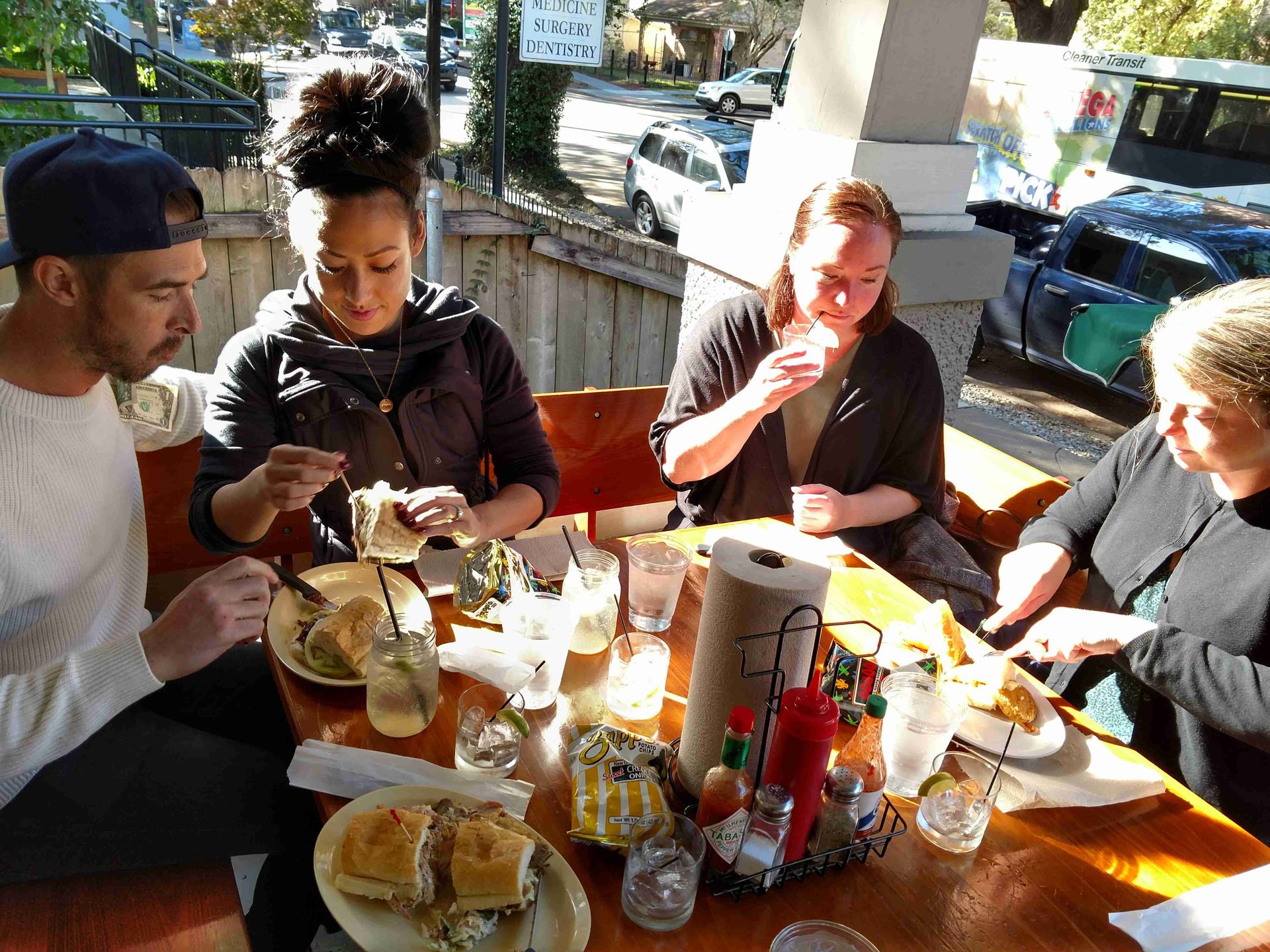 Sunlit outdoor cafe patio scene with four people in a group eating sandwiches and chips at a wooden table, mason-jar drinks, condiments, and parked cars on the street in the background.