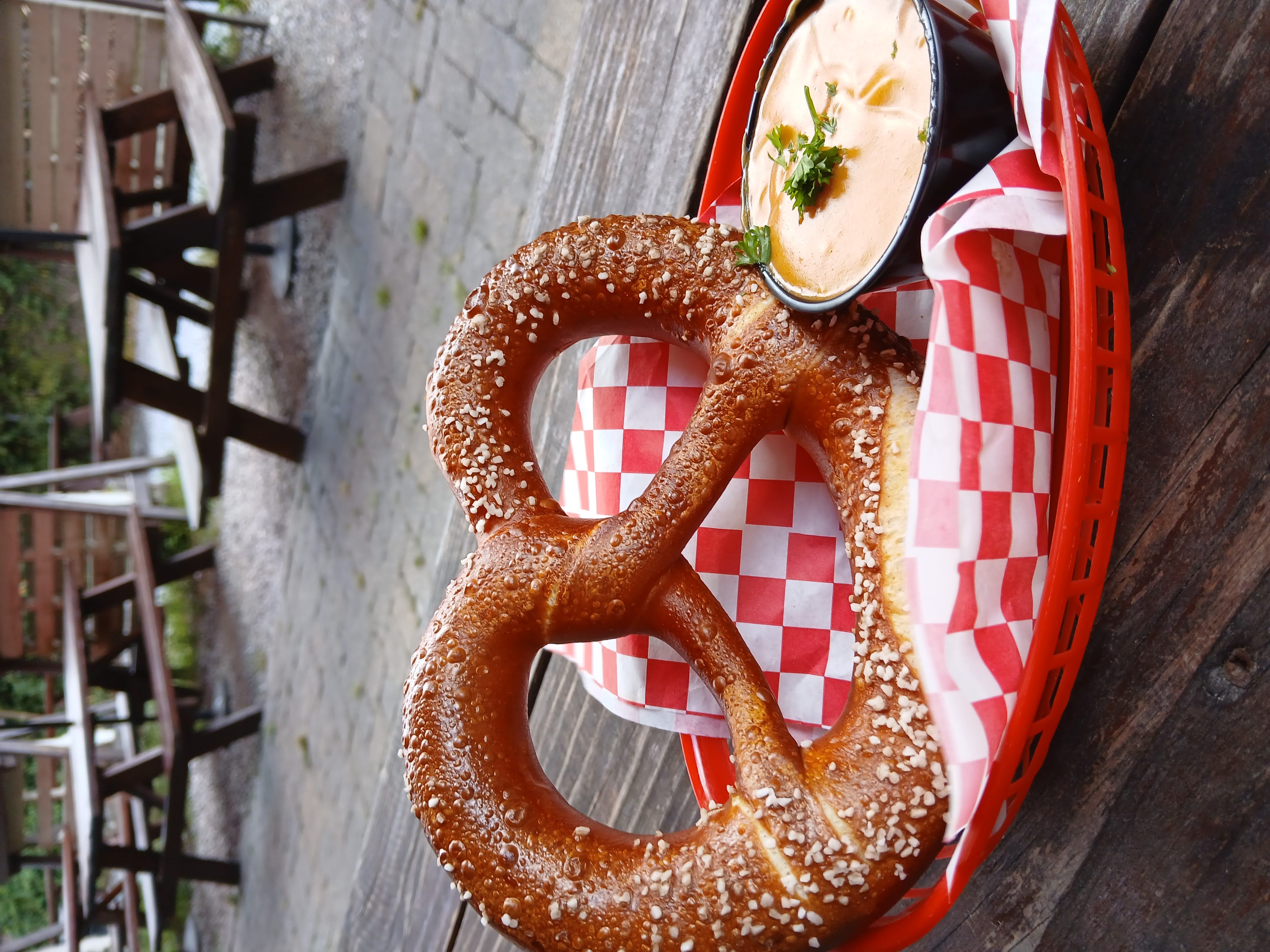 Salted soft pretzel in a red basket on red-and-white checkered paper with a cup of creamy beer-cheese dip topped with parsley, sitting on a wooden table with outdoor picnic tables in the background.