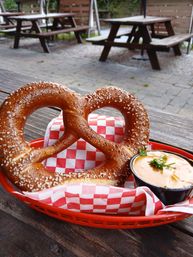 Salted soft pretzel in a red basket on red-and-white checkered paper with a cup of creamy beer-cheese dip topped with parsley, sitting on a wooden table with outdoor picnic tables in the background.