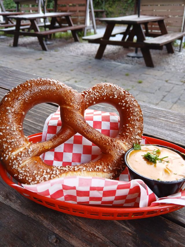 Salted soft pretzel in a red basket on red-and-white checkered paper with a cup of creamy beer-cheese dip topped with parsley, sitting on a wooden table with outdoor picnic tables in the background.