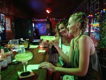 Two friends clinking salt-rimmed margaritas at a festive, neon-lit bar counter with colorful string lights and tinsel, capturing a lively nightlife moment.