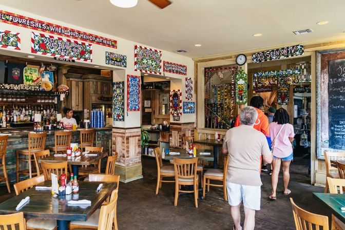 Casual restaurant interior with wooden tables and bar seating, patrons queuing at the counter, colorful folk-art signs and a chalkboard menu.