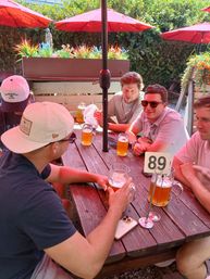 Group of friends enjoying large beer mugs at a wooden picnic table on a sunny outdoor patio with red umbrellas, planters, and table number 89.