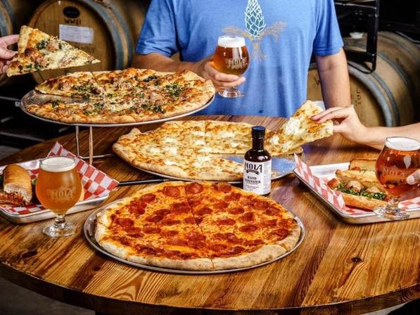 Three large pizzas (pepperoni, white, and herb) on metal trays with craft beer glasses and sandwich sides on a rustic wooden table, hands reaching for slices in a casual taproom setting.