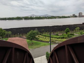 View from a rusty metal observation platform over a riverfront park with brick walkways and planted gardens, wide muddy river and distant steel truss bridge under an overcast sky.