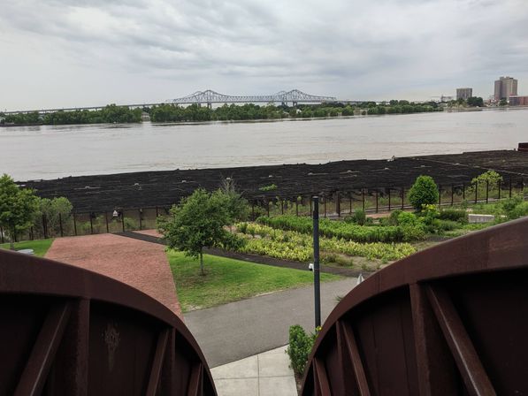 View from a rusty metal observation platform over a riverfront park with brick walkways and planted gardens, wide muddy river and distant steel truss bridge under an overcast sky.