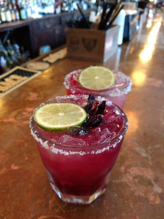 Two vibrant purple hibiscus margaritas with salted rims, lime slices and crushed ice on a copper bar top — craft cocktails at a lively bar.
