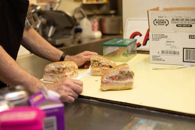 Three wrapped deli sandwich halves on a shop counter, layered with meats, cheese and chopped vegetables, with a worker's hands nearby in a commercial kitchen.