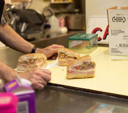 Three wrapped deli sandwich halves on a shop counter, layered with meats, cheese and chopped vegetables, with a worker's hands nearby in a commercial kitchen.