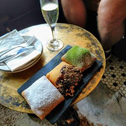 Three square beignets on a slate board — one dusted with powdered sugar, one topped with matcha, and one with chocolate and crunchy crumble — served with a flute of sparkling wine and stacked plates on a brass café table.