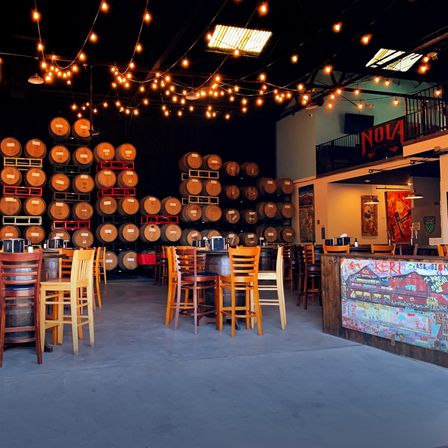 Warehouse-style brewery taproom with stacked oak barrels on the back wall, wooden high-top tables and chairs, warm string lights overhead, and a colorful mural-accented bar.